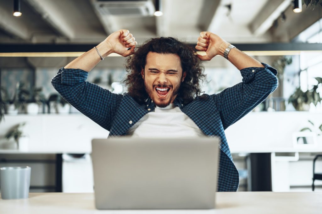 Excited overjoyed young businessman looking at laptop screen happy to win or recieved good news