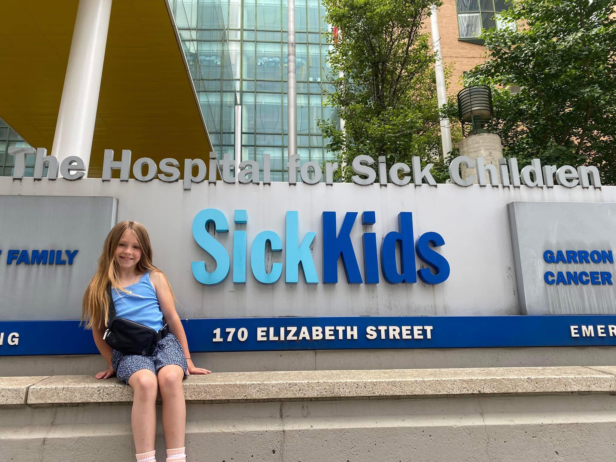 Girl sits on concrete steps in front of The Hospital for Sick Children sign, with big blue 'SickKids' letters and a glass-fronted building behind her outside the hospital entrance on Elizabeth Street.