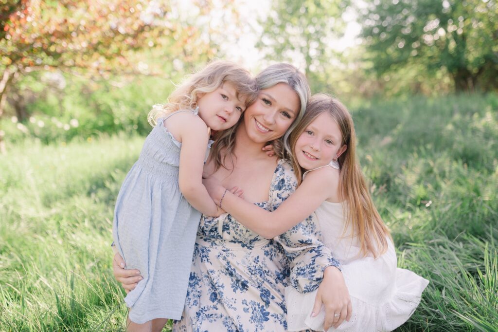 Mother hugs her two young daughters in a sunlit meadow, all smiling at the camera with green grass around them.
