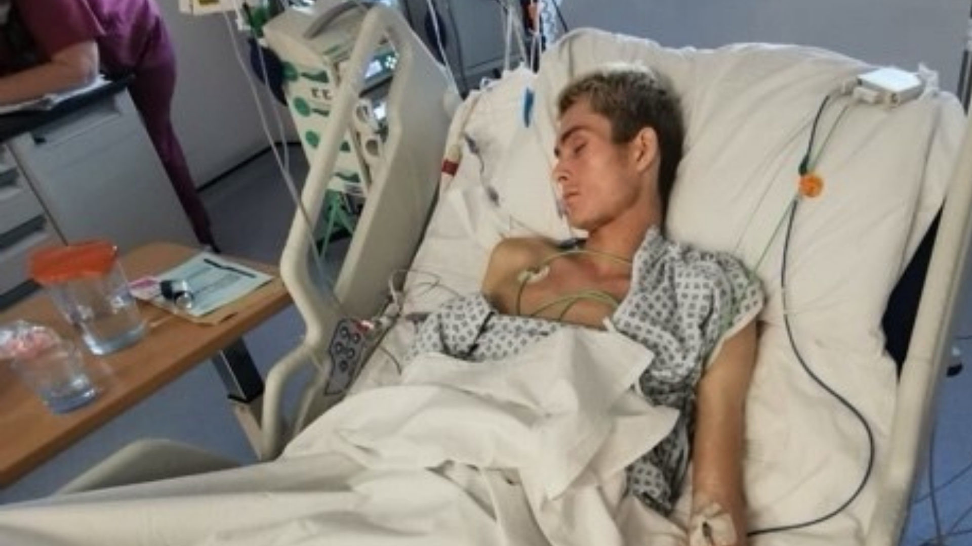 Patient lying in a hospital bed connected to monitors and tubes, resting with eyes closed.