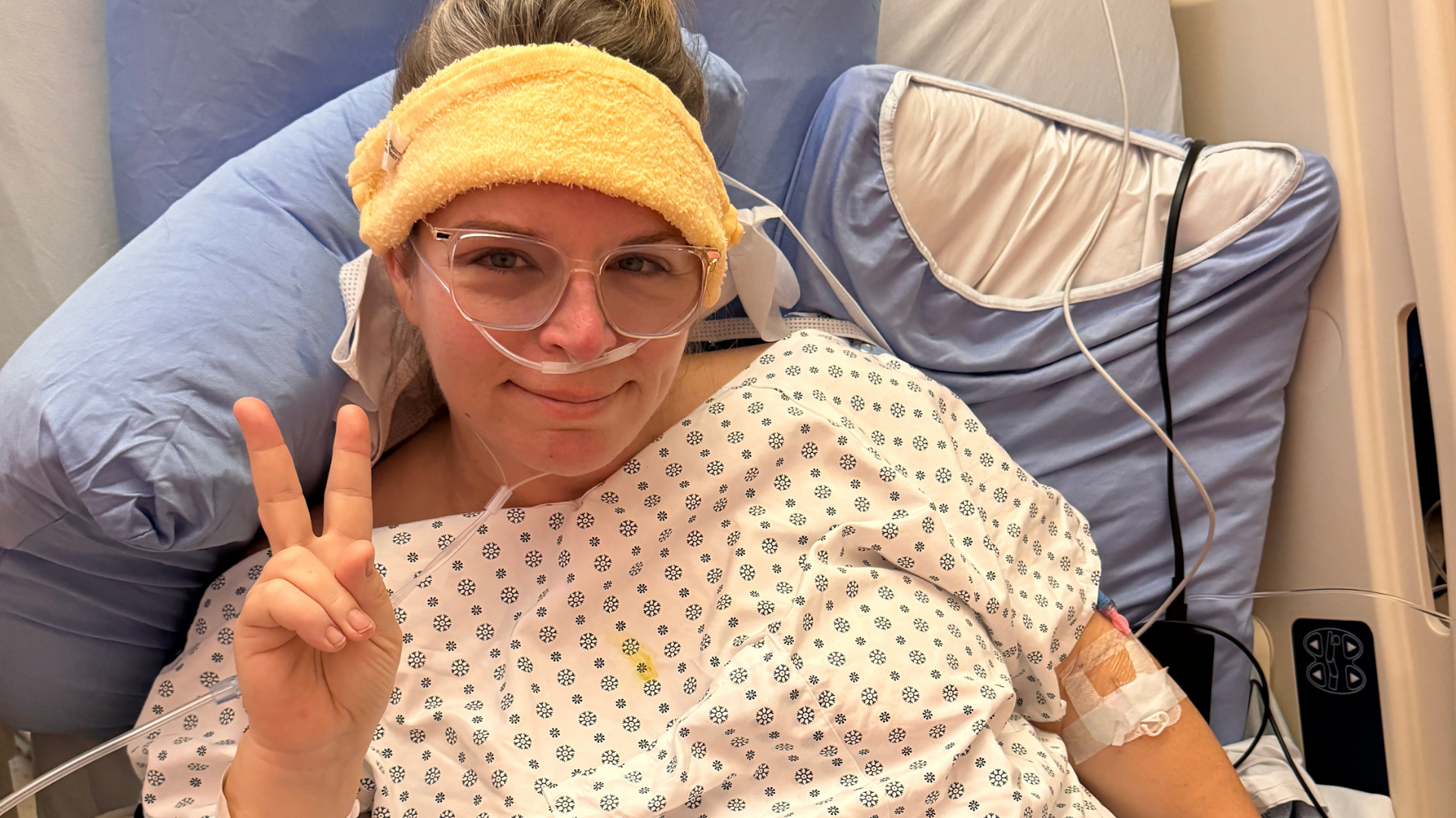 Patient in a hospital bed with a yellow head wrap, smiling and making a peace sign, oxygen tubes in nose.